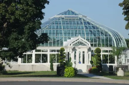 Ici, un bâtiment muni d’une voûte de verre comme toit occupe le centre de la photographie. La porte blanche du bâtiment est entourée d’arbustes verts. À travers le verre, on voit des plantes vertes de différentes tailles. Au premier plan, des branches d’arbres feuillus entrecoupent la photographie.
