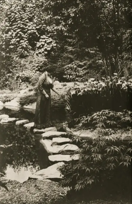 Sur cette photographie en noir et blanc, Jennie Butchart se promène dans son jardin. Elle se tient sur l’un des pavés du sentier qui traverse le ruisseau. À l’arrière-plan, on aperçoit des arbres feuillus. Au premier plan, des arbustes de différentes tailles bordent le sentier et le ruisseau.