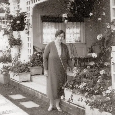 Jennie Butchart se trouve au centre de la première photographie en noir et blanc. Elle pose devant sa résidence où l’on voit plusieurs jardinières. Quelques-unes sont suspendues à un treillis; d’autres embellissent le sol. Elles sont toutes garnies de plantes de tailles et de floraisons distinctes.